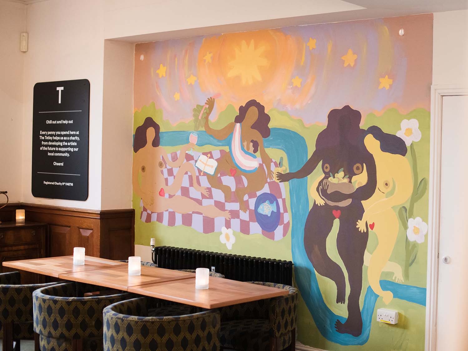 A brightly coloured mural with dancing naked people around a picnic blanket. The mural is painted on a wall in The Tetley's Bar & Kitchen