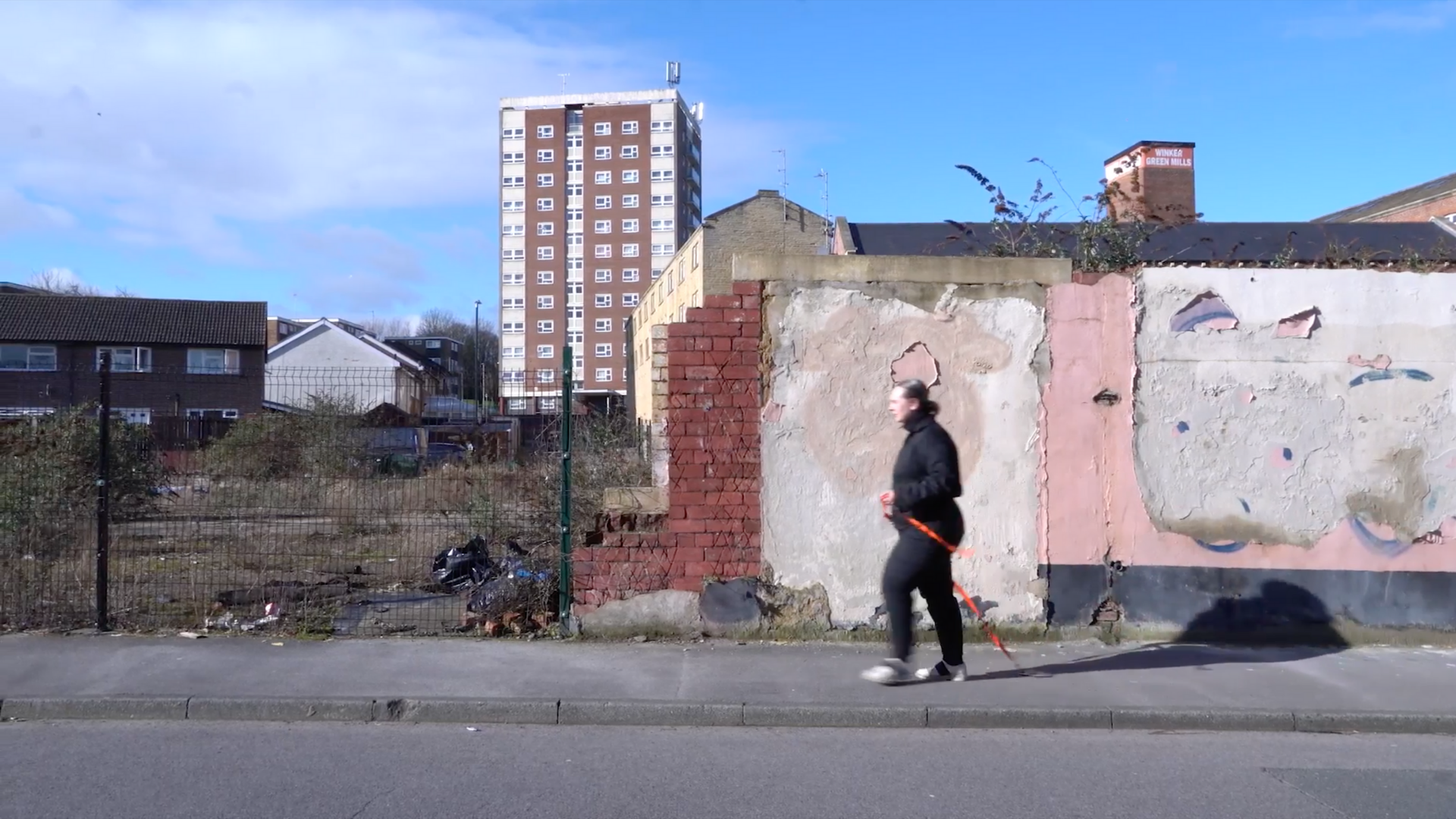 City scene, pavement in the foreground, lined with a patchwork concrete and brick wall, a tall green wire fence blocking off an overgrown concrete slab with bin bags. Houses and an 11-storey high-rise are in the background. A person in a black jumpsuit is in the centre frame, walking across the pavement, pulling a neon orange ratchet strap attached to a small metal plate which is being dragged along the pavement.