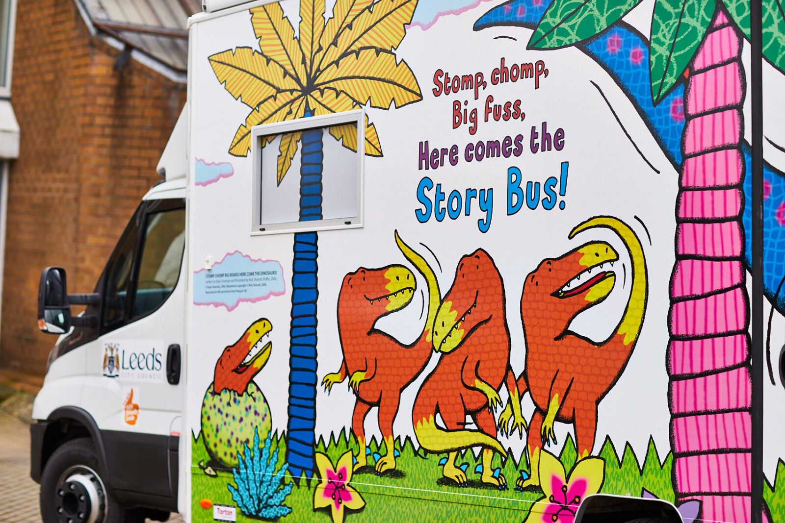 A white bus with illustrated dinosaurs. It says 'stomp chomp, big fuss. Here comes the Story Bus!'