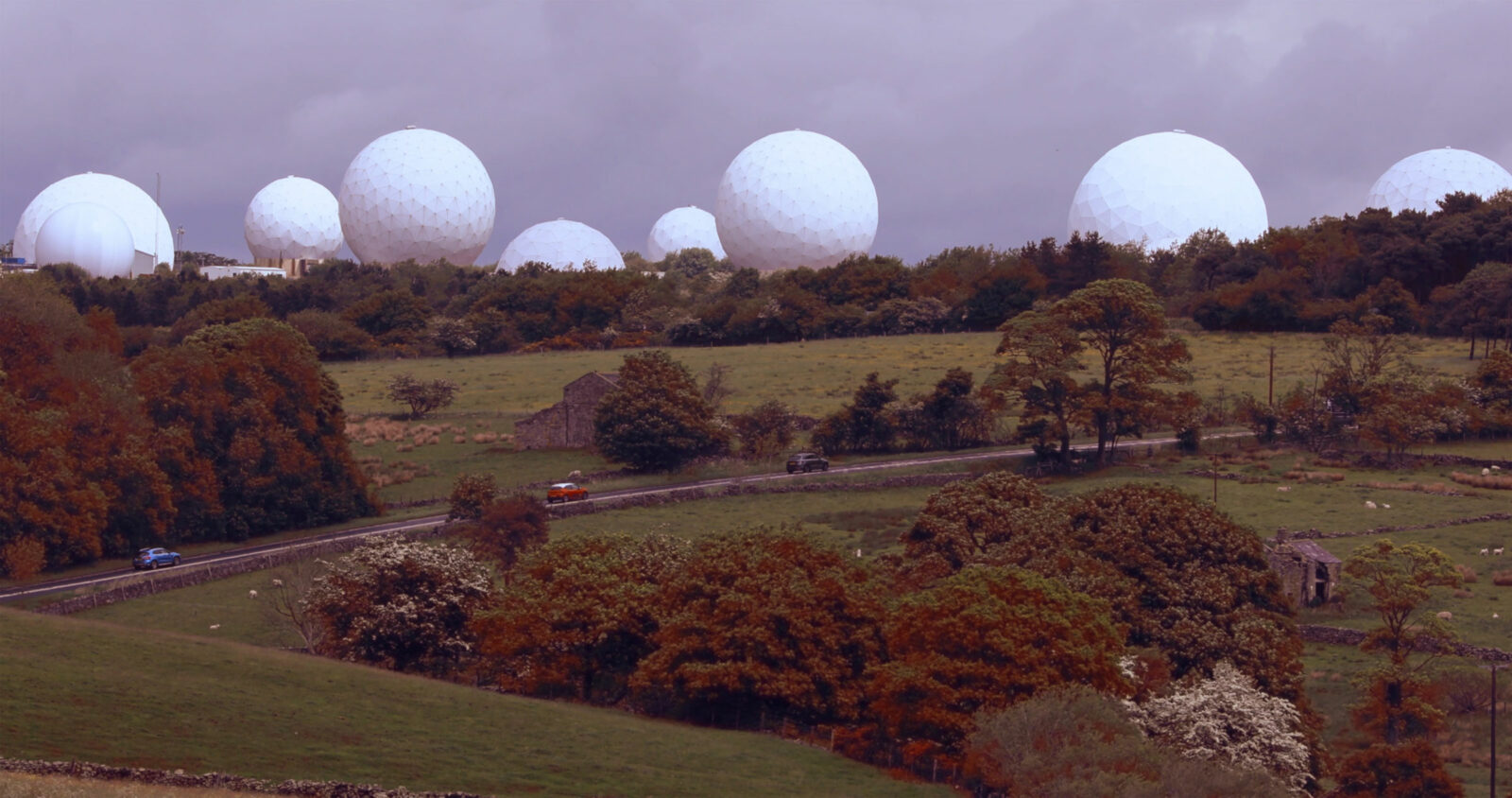 a Northern English landscape with rolling hills is intersected by drystone walls, stone houses, and trees in sepia-like tones of green, brown and burnt yellow. Looming large on the horizon above the more tranquil valley are 27 white surveillance radomes. The sky is a flat grey behind these huge geodesic domes.